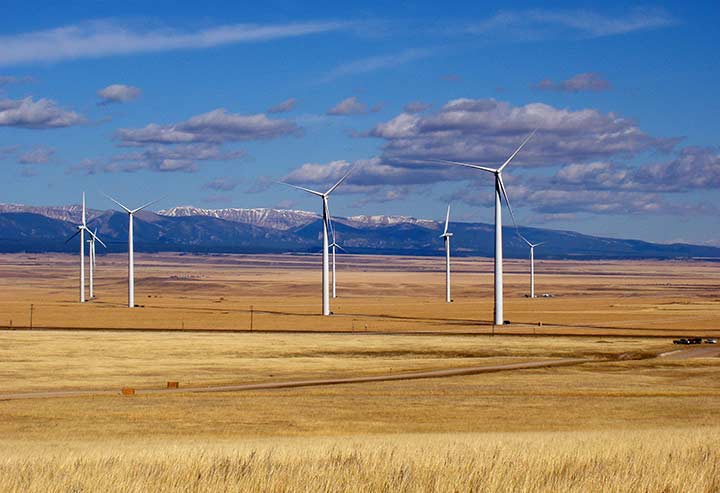 Multiple wind turbines on a flat landscape with mountains in the back.