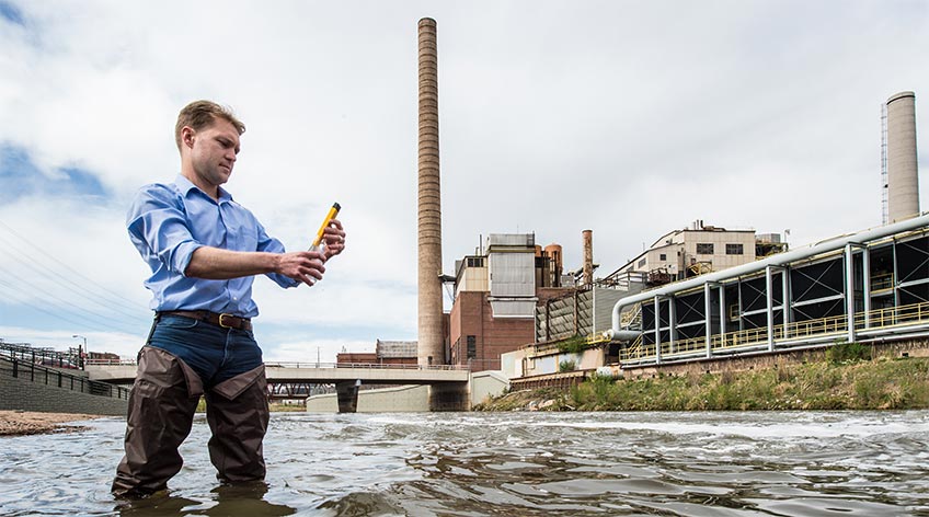 Photo of a man standing in water looking at a water sample in front of a hydropower plant.