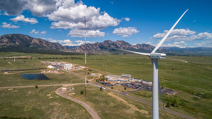A wind turbine stands in the foreground with roads and buildings below. A mountain range spans the background.