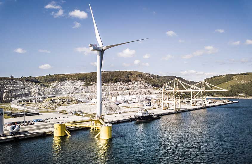 A wind turbine erected on one column of a three-sided structure floating in the ocean next to a dock.