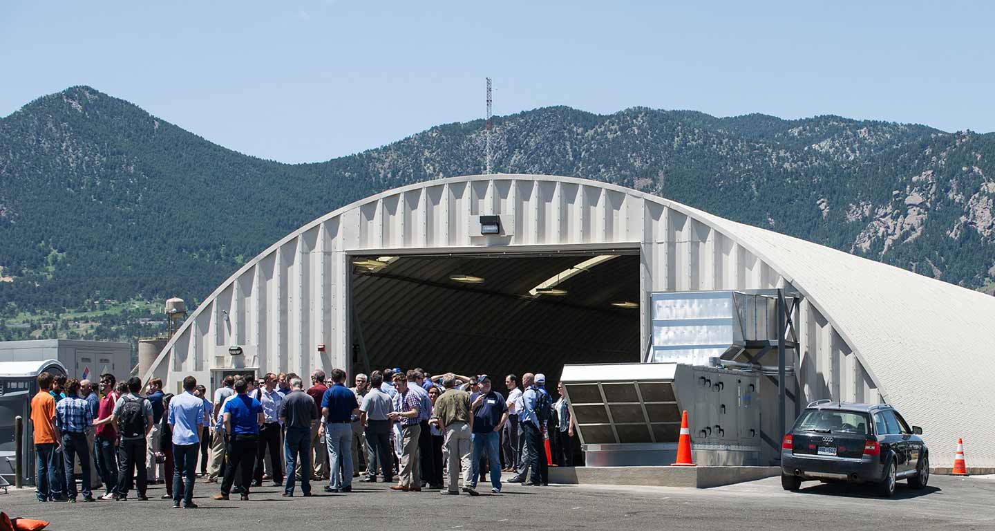 A large group of people stand outside of a white, half-cylindrical facility with mountains in the background.