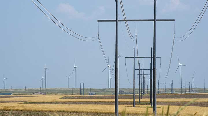 A photo of a transmission power line in a brown field with wind turbines in the background.