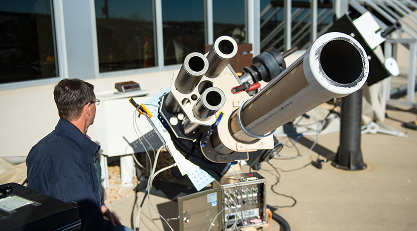 Photo of a man sitting outside next to large, silver collimating tubes for testing solar cells