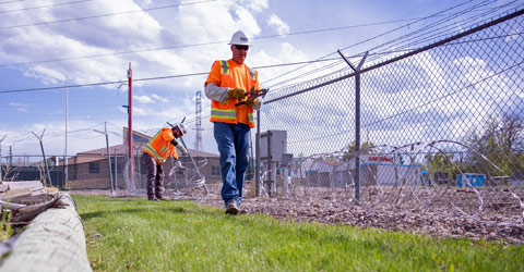 Two people in bright vests and hard hats work near a wire fence.