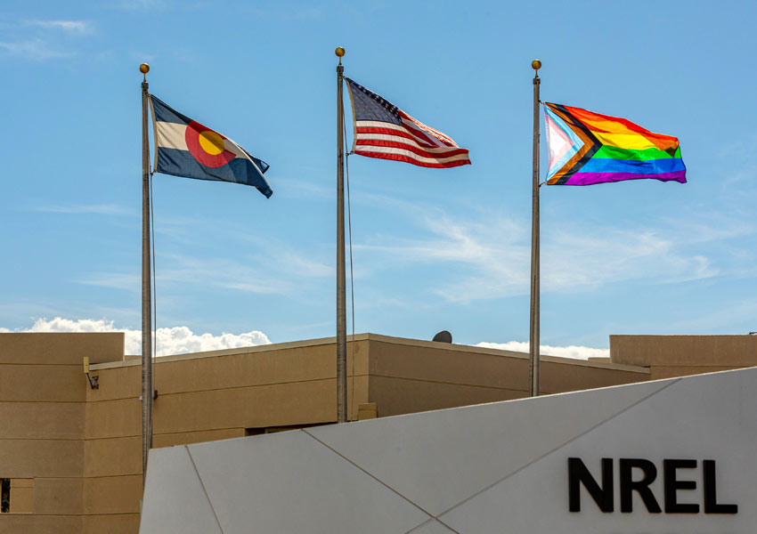 The Colorado state flag, the American flag, and the Progress Pride Flag flying, with a sign that says "NREL" in the foreground.