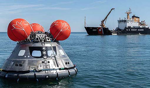 A NASA capsule floats in the ocean with a Coast Guard ship in the background.