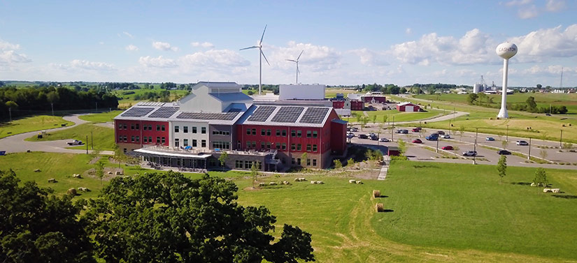 Photo of a building in a rural setting. The building has solar panels and medium-size wind turbines in the background.