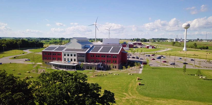 Photo of a building in a rural setting. The building has solar panels and medium-size wind turbines in the background.