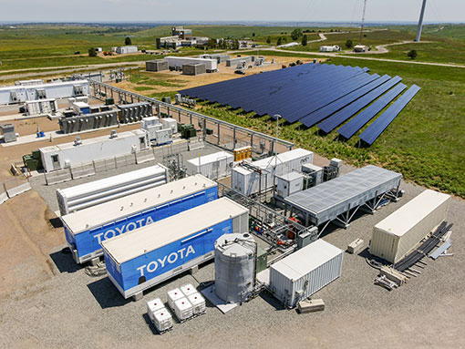 Aerial shot of energy storage containers outside with solar array and wind turbine in the background.