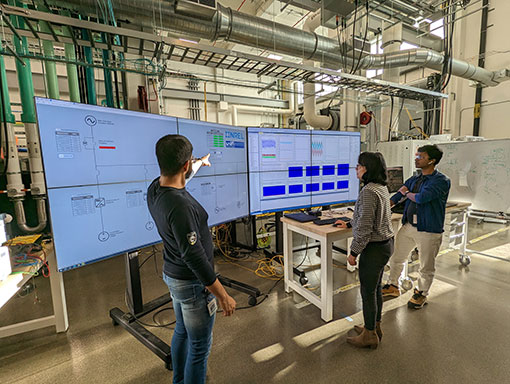 Three people stand inside lab pointing and looking at data on large display screens