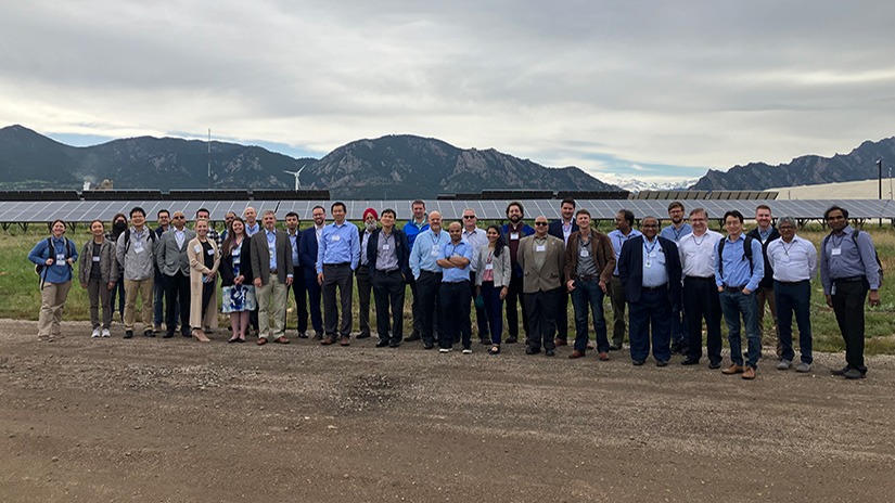 Group of people stand in front of solar panels, wind turbines, and mountains.