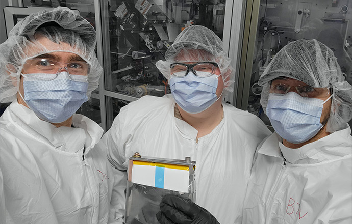 Three people wearing personal protective equipment hold up a large battery to display the electrodes