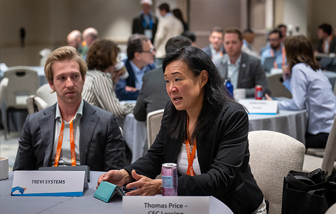 Two people sitting and talking at a table in a large conference hall full of other tables with people