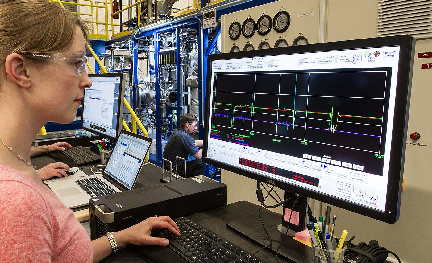 Woman in front of monitor displaying line graph in industrial facility; other workers in background.