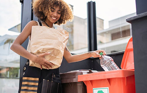 A woman holding a paper bag places plastic bottles into a recycling bin.