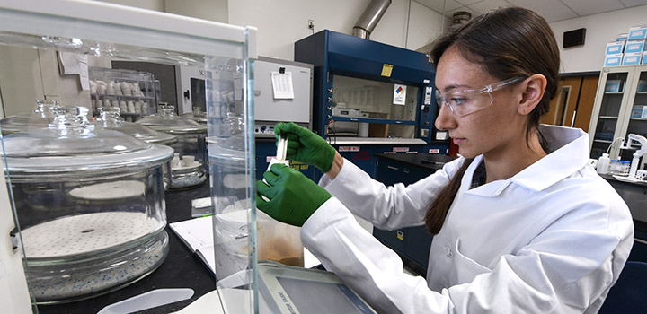  A woman in safety glasses, rubber gloves, and lab coat measures a sample in a laboratory.
