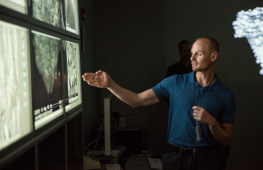 Man pointing to a series of large computer monitors showing molecular images.