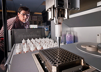 Photo of a male scientist wearing safety glasses in a lab watching a robot dispensing samples of powdered biomass into a reactor plate, part of the high-throughput biomass recalcitrance pipeline to analyze the way potential biofuels feedstocks give up their sugars.