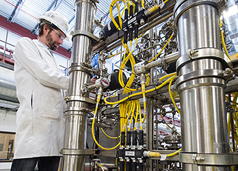 Photo of a man in a hardhat and safety glasses in front of shiny metal pipes and yellow tubes, working on a cross flow membrane filter in the industrial plant setting.