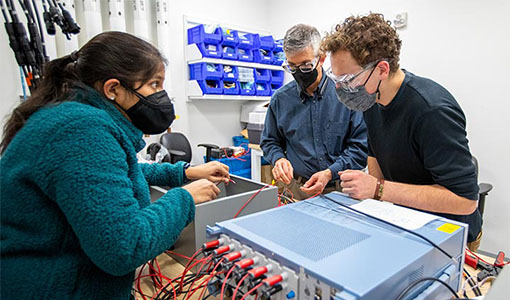 Three people work together in a laboratory connecting wires to metal boxes, preparing the equipment for testing.