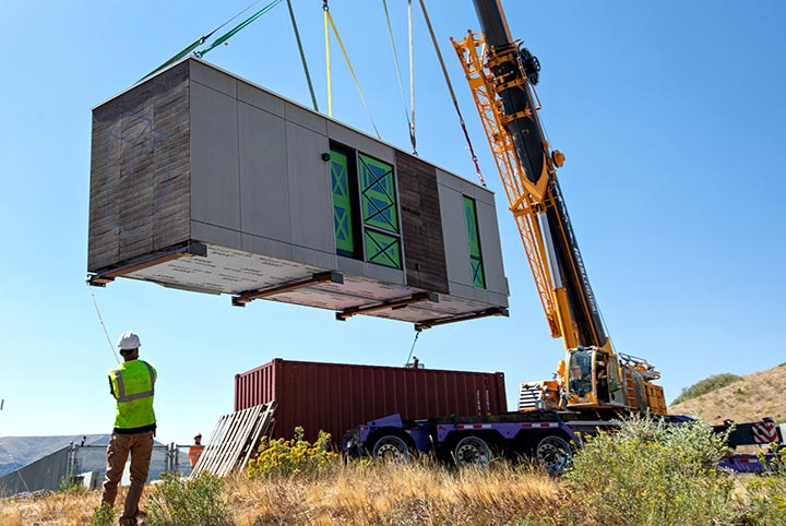 A crane places a small, pre-fabricated apartment unit on a grassy site.