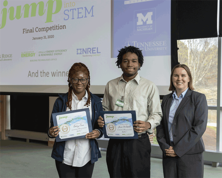 Photo of two students, holding winner certificates and standing next to an adult in business attire.