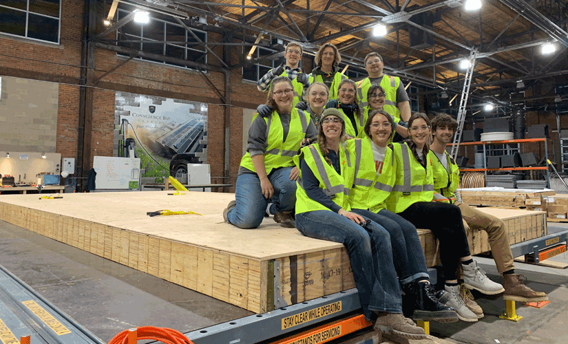 Photo of 11 students, wearing safety glasses and vests, in a warehouse with construction materials.