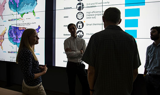 Four researchers conversing in front of two large data visualizations with U.S. maps and charts.