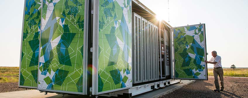 Photo of a man closing the door of a large, multicolored metal box that is roughly the size of a semi-truck trailer. Inside the box are batteries that make up a 1-megawatt battery energy storage system.