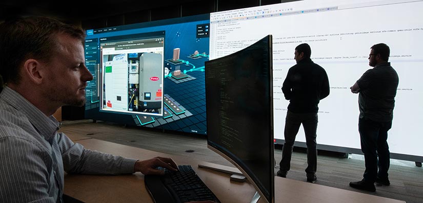 Photo of a researcher on the computer sitting at a desk and two researchers standing in front of a large monitor emulating a real-time cyberattack.