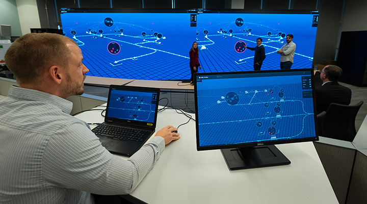 Photo of a researcher on the computer sitting at a desk and a researchers standing in front of a large monitor emulating a real-time cyberattack.