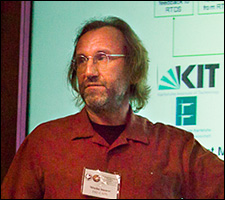 A photo of a man with a beard wearing a red shirt pointing to a PowerPoint presentation behind him.