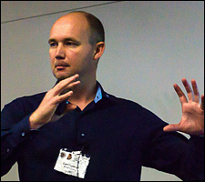 A photo of a man with a shaved head wearing a navy blue shirt gesturing with his hands.