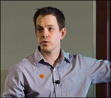 A photo of a young man wearing a stripe shirt standing in front of a PowerPoint presentation.