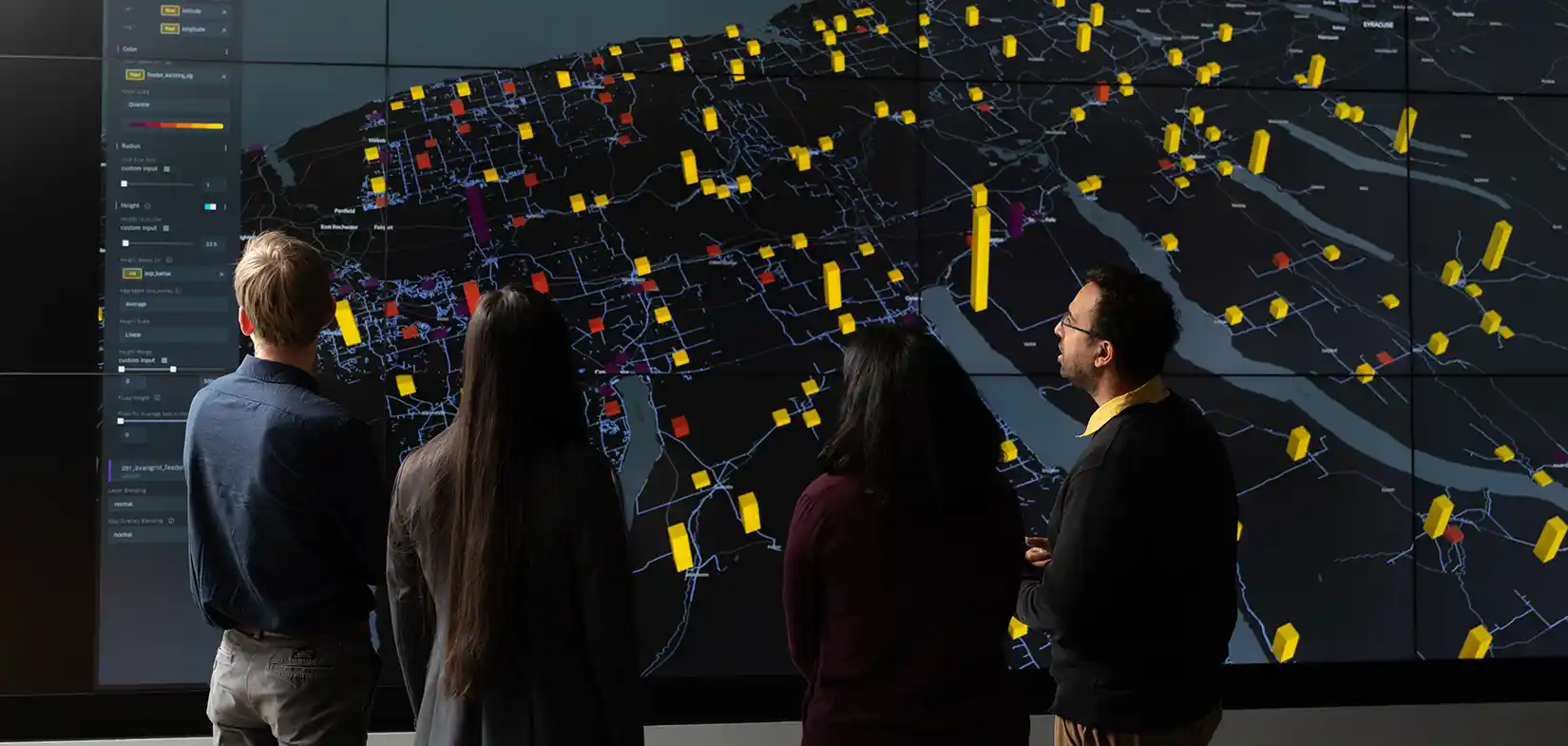 Four people stand in front of large display screen looking at grid data.