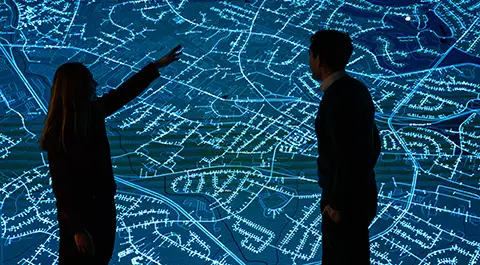 Two people point at grid data on large display screen.