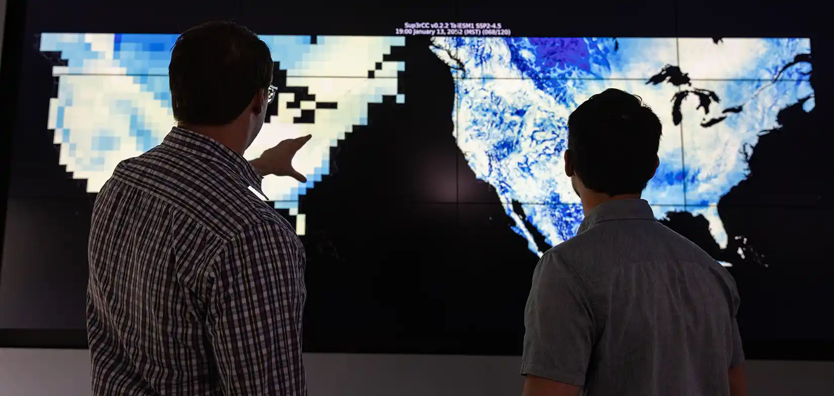 Two people point to maps of the United States on a large data display screen.