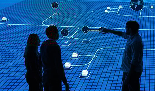 impacts-74769 Three people stand in front of an point at large screen with grid data on display.