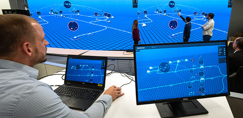 industry-cyberrange Person navigates laptop while screen displays on a large screen where other stand in front and point.