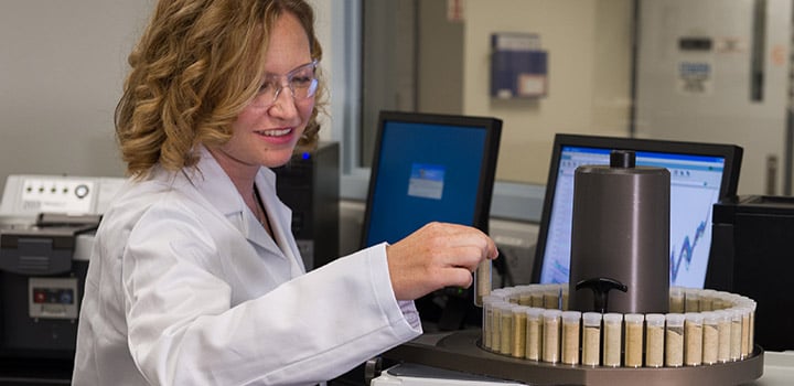A woman in a lab coat and safety glasses looks at a vial in a laboratory.