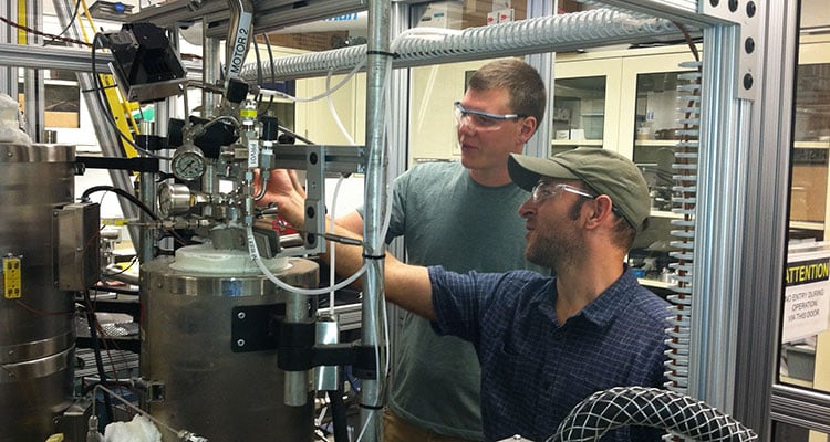 tcuf-28346 Photo of two engineers in safety glasses working on a reactor system with metal tanks, tubing, pipes, and wires. The system is used for in situ and ex situ catalytic fast pyrolysis to generate bio-oil for compositional analysis.