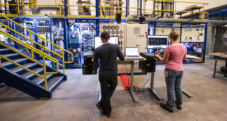 tcuf-33048 Photo of two engineers facing a large, multi-level Davison circulating riser, with laptops on tall tables. The riser features colorful scaffolding, pipes, tubes, and control panels with dials and monitors.