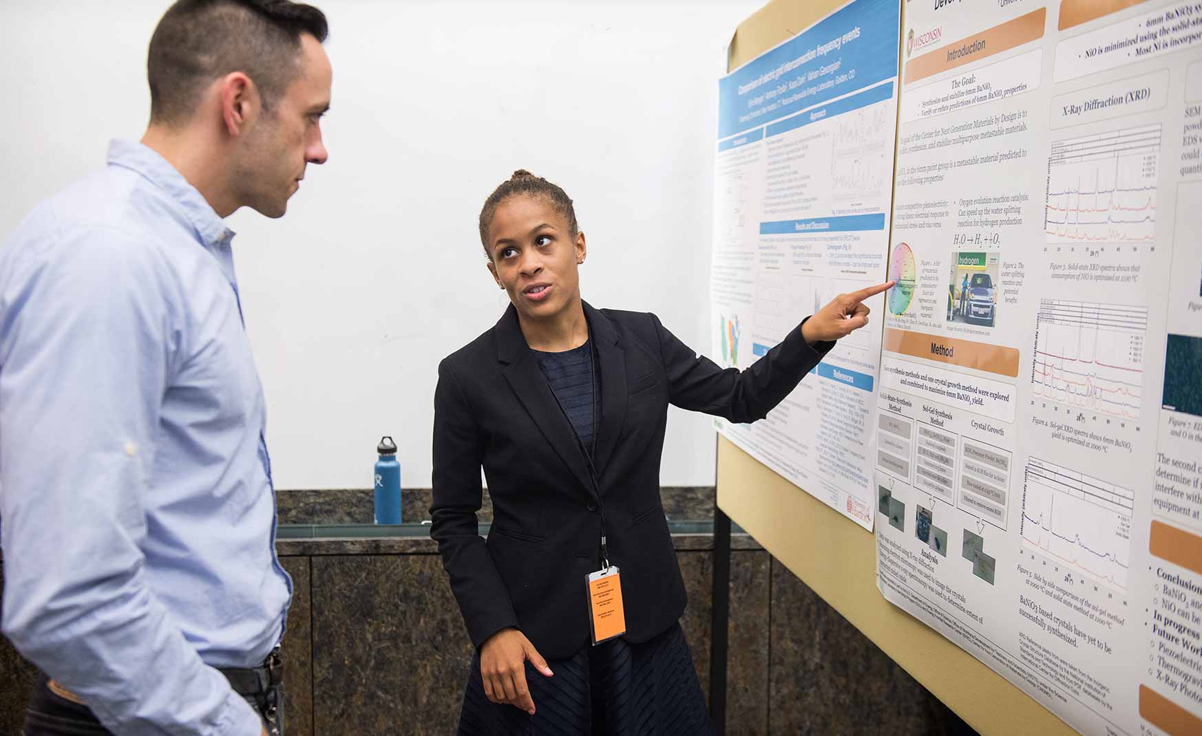 cci-riley-whitehead One person talks and points at a poster hanging on a board while another listens.