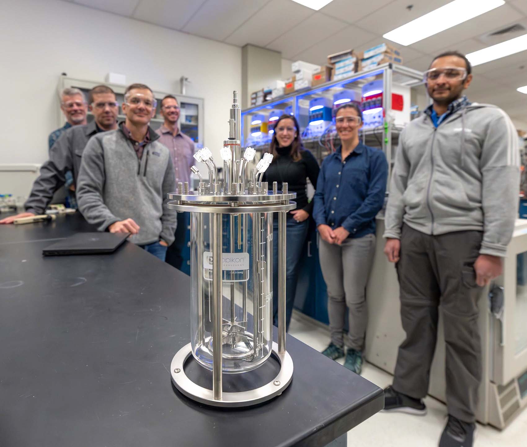 reactor-team A group of seven individuals stands in a laboratory behind a cylindrical device.