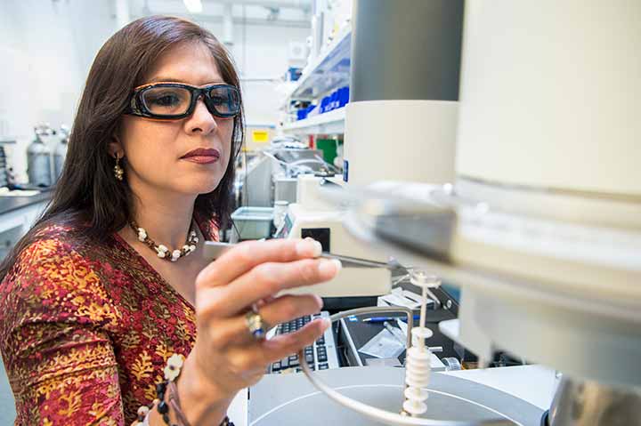 201909-facilities-45088 A woman in safety glasses reaches toward a piece of laboratoy equipment.