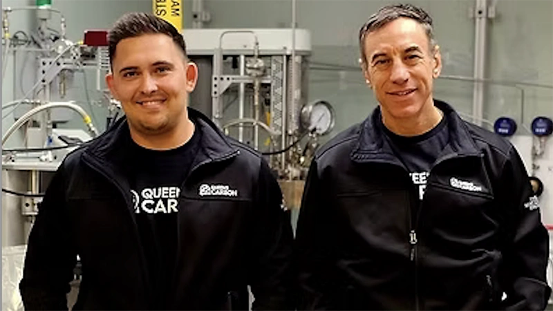 Two people in black jackets stand in front of industrial equipment.