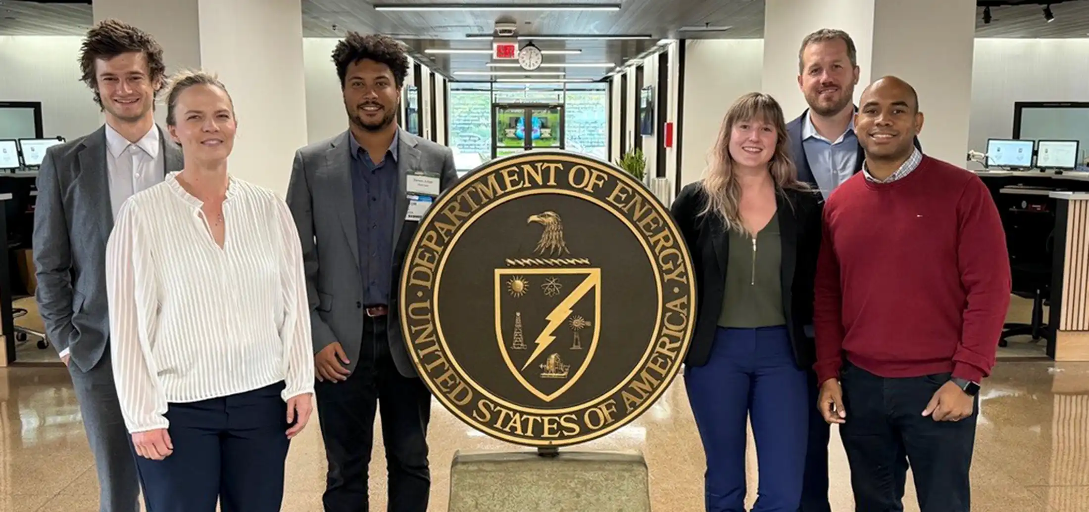 Six people stand beside a large circular metal seal reading ‘Department of Energy United States of America’.