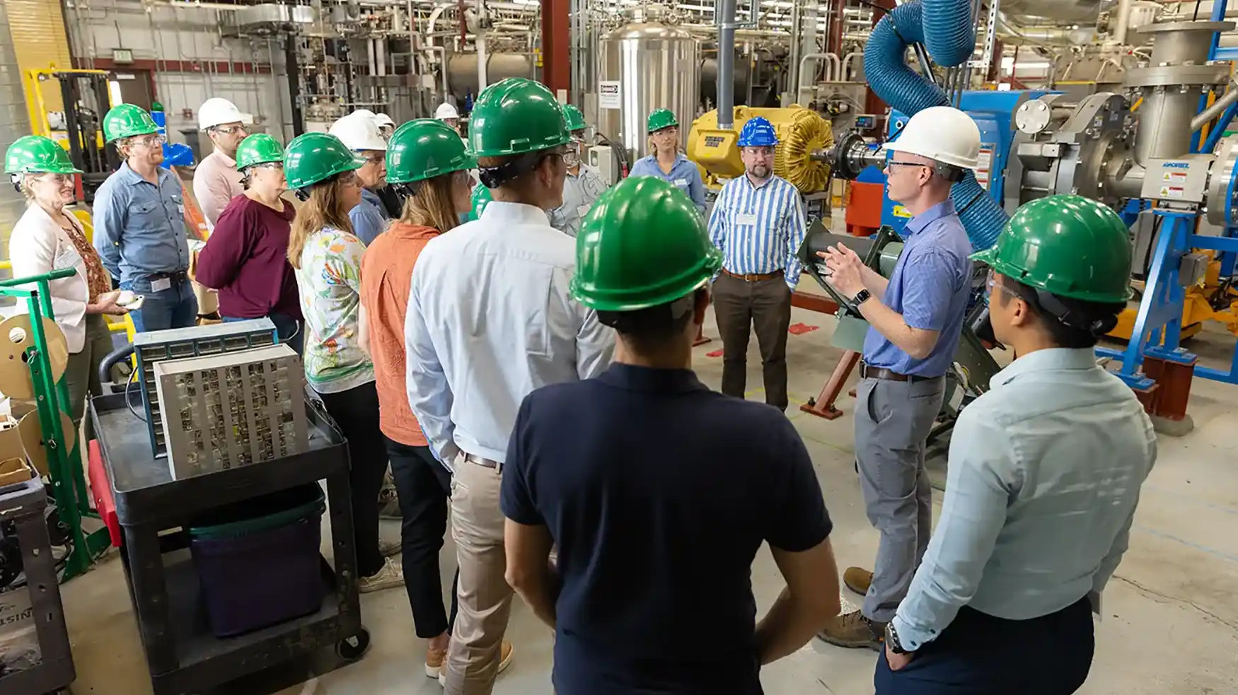 A group of people wearing hard hats are gathered around a tour guide speaking to them inside a large research lab facility.