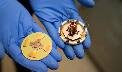 Two gloved hands hold a small octagonal power module and a corresponding attachment layer.