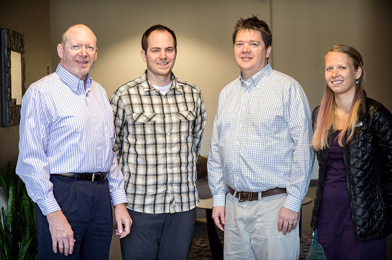 Three men and a woman stand and smile in a room.